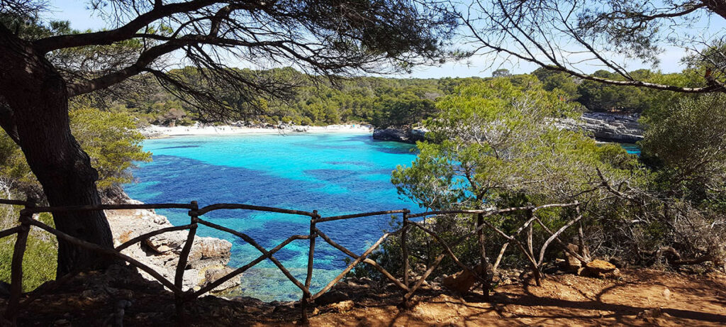 Stunning view of Cala Turqueta in Mallorca, featuring crystal-clear turquoise waters, a secluded white sandy beach surrounded by rugged cliffs and lush green forest.