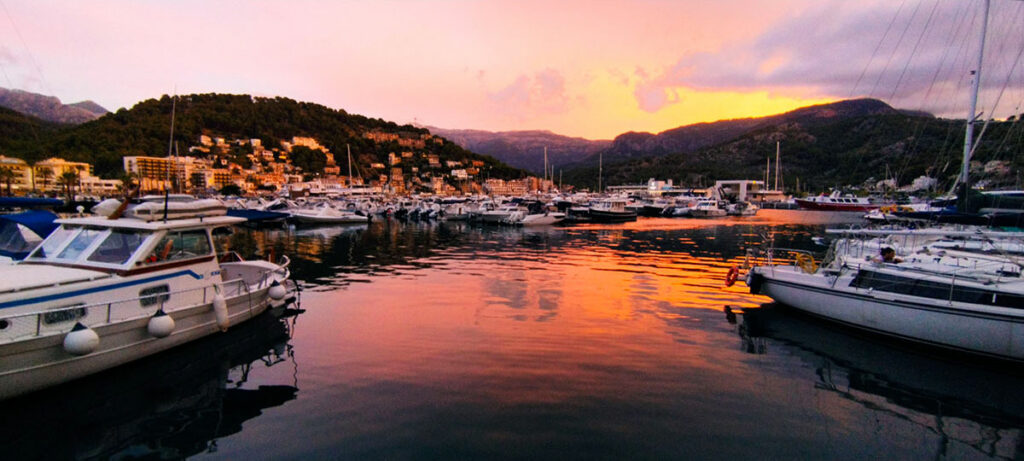 Sunset at Sollers marina, Mallorca: A breathtaking scene capturing boats moored in the serene marina with a stunning backdrop of orange and pink skies delicately mirroring on the calm waters, surrounded by lush, rolling hills.