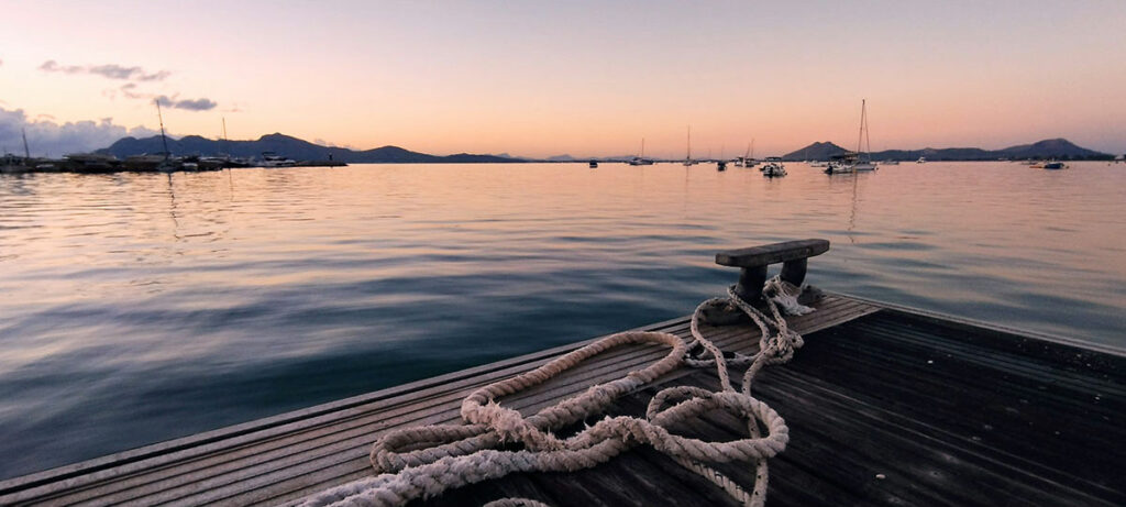 Scenic sunrise view at the Pollença Pier, featuring the serene and golden-lit waters of the bay, with the picturesque pier extending into the calm sea under a pastel-colored sky.