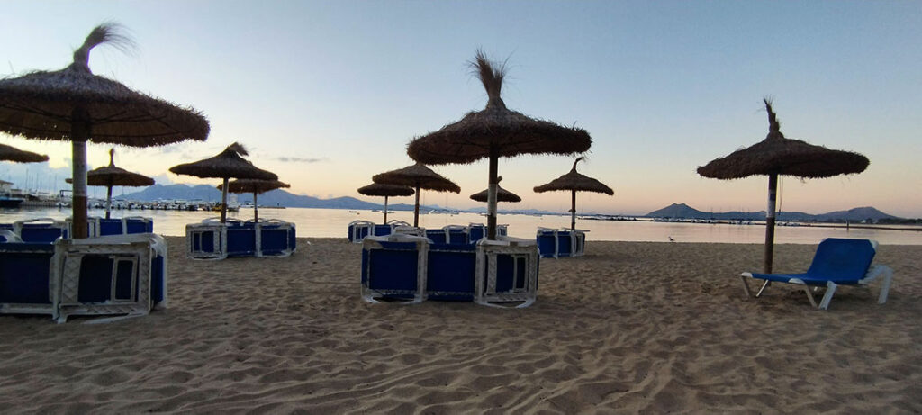 Scenic sunrise view at the Pollença playa, featuring the serene and golden-lit waters of the bay, with the picturesque pier extending into the calm sea under a pastel-colored sky.