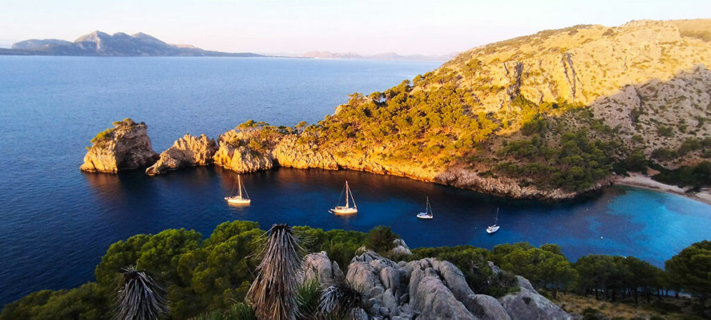 Stunning aerial view of Cala Murta at sunrise, showcasing the serene bay with crystal-clear waters and pristine sandy beach, surrounded by rugged cliffs and lush greenery under a colorful sky.